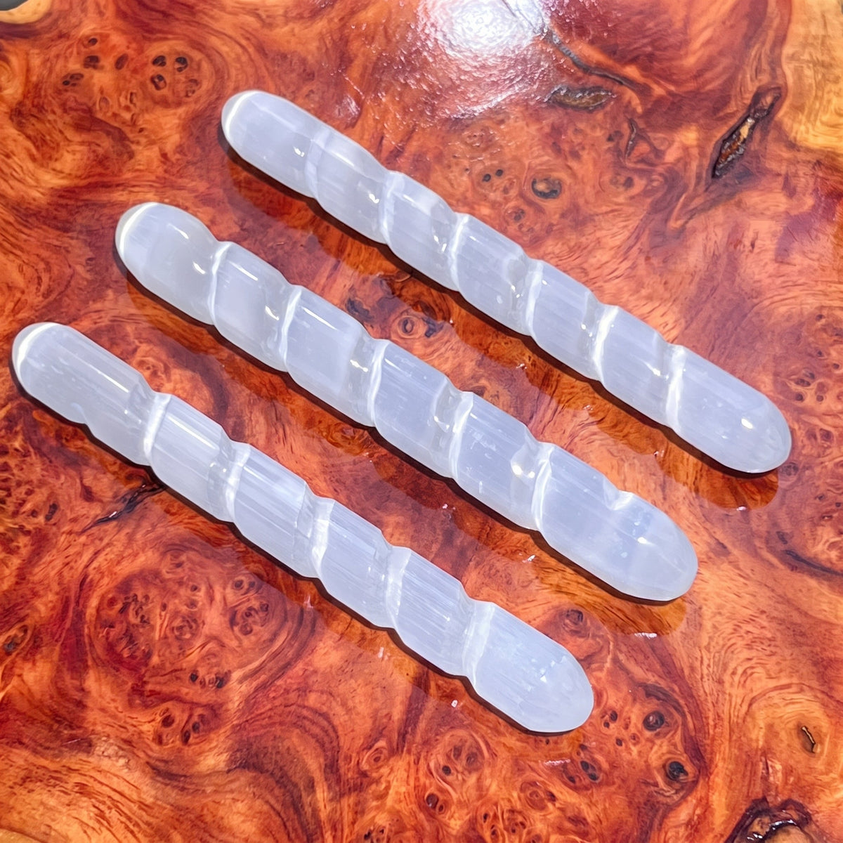 Three white selenite satin spar wands with carved spiral grooves arranged on reddish patterned wood.
