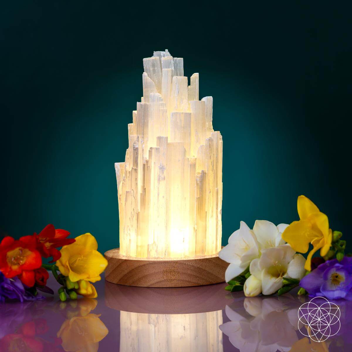 Glowing white selenite tower lamp on a round walnut base surrounded by colorful flowers against a teal background.
