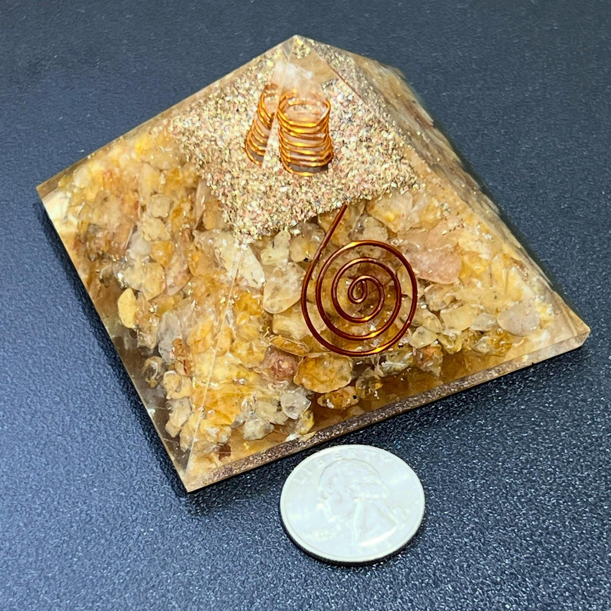 Angled view of a yellow quartz resin pyramid with embedded crystal chips and spiral details beside a coin for size comparison.